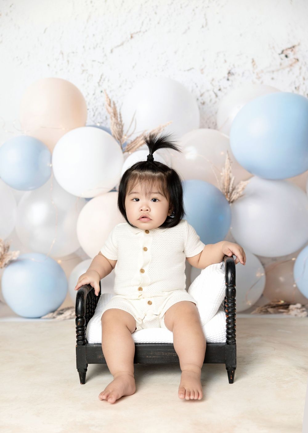 An adorable baby girl sits in a mini chair surrounded by pastel balloons in a birthday photo session.