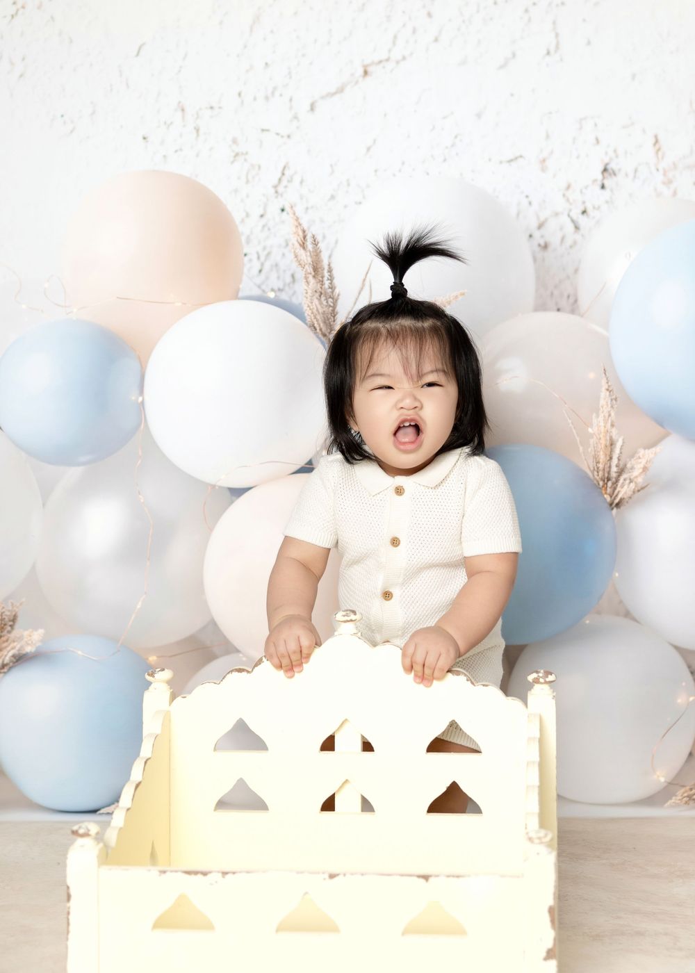 Laughing baby in cream outfit sits in white crate surrounded by pastel blue and white balloons.