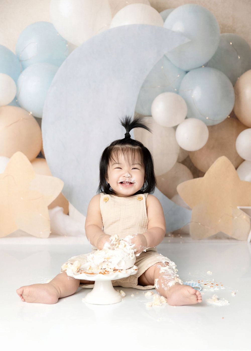Smiling baby girl enjoys first birthday cake smash with blue balloons and moon backdrop.