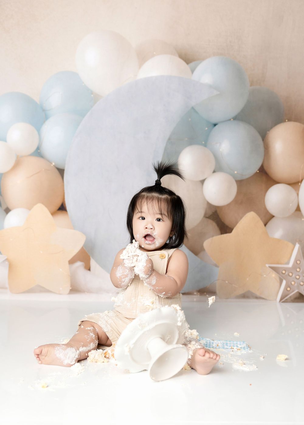 A baby girl enjoys her first birthday cake smash surrounded by blue, white, and beige balloons and star decorations.