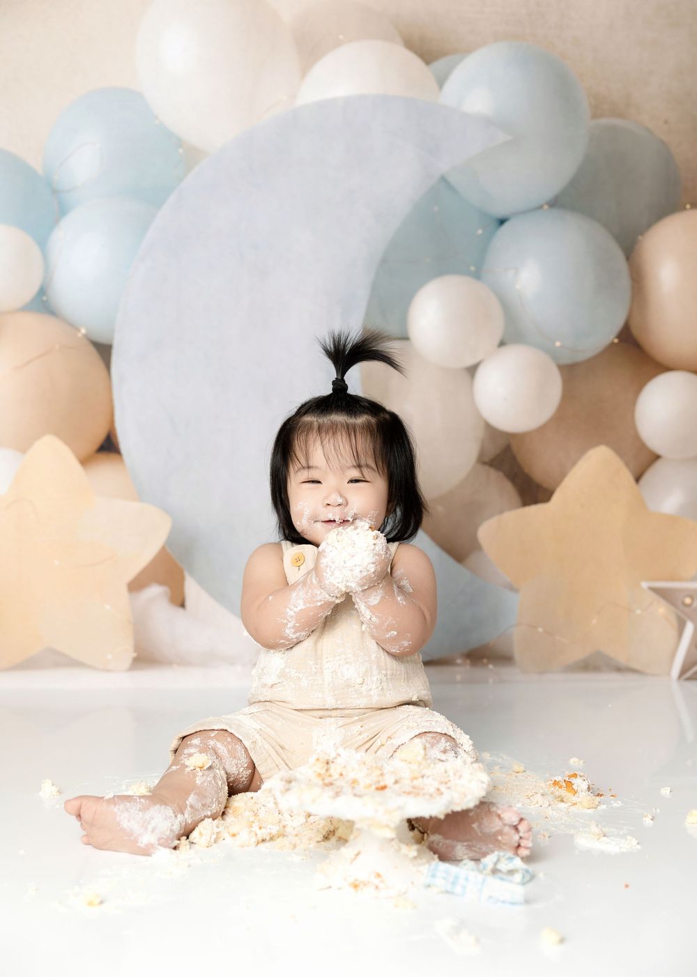 Happy toddler smashes first birthday cake surrounded by moon and star decorations with blue balloons.