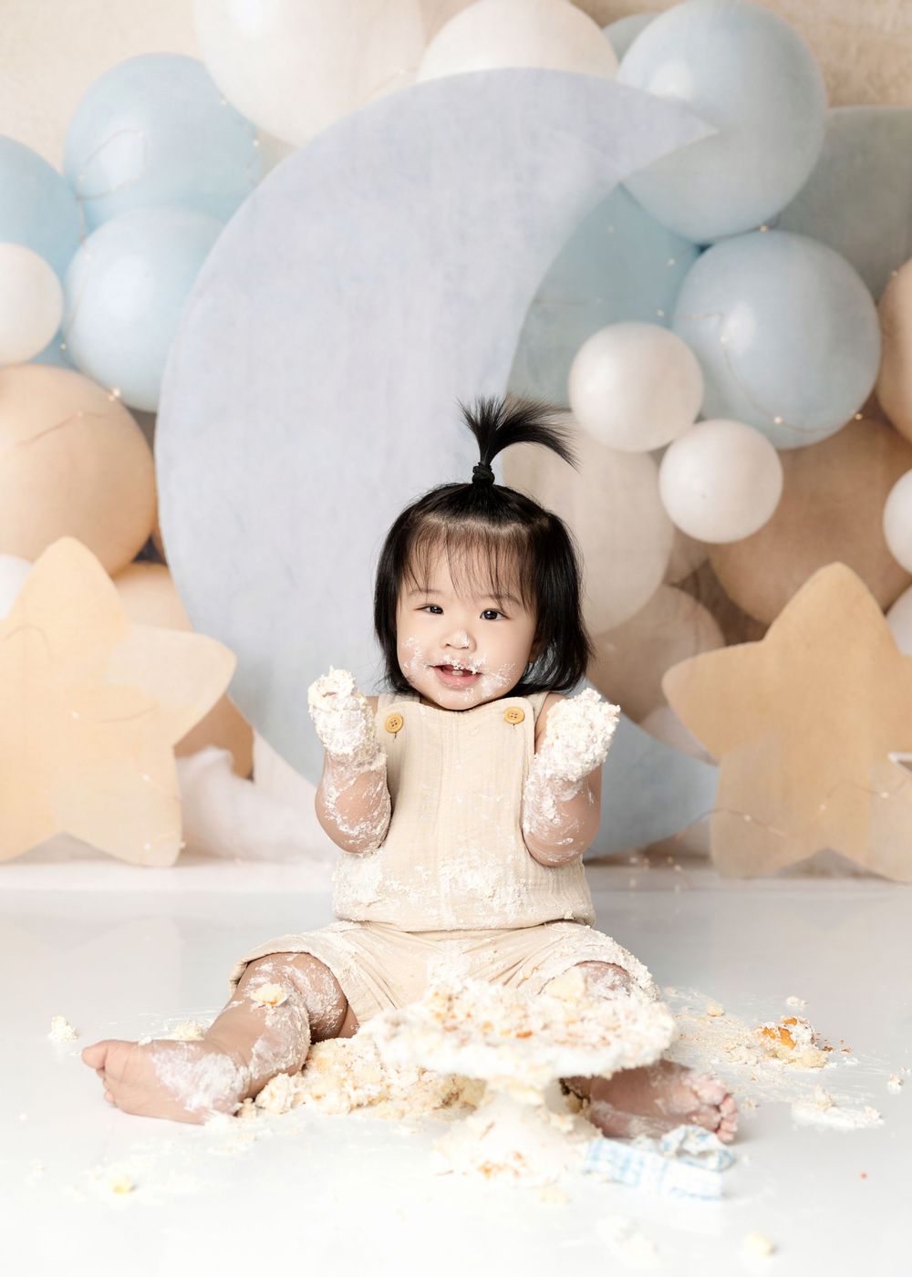 A joyful baby girl smashes her first birthday cake surrounded by pastel balloons and star decorations in a studio.