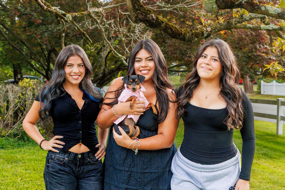 Three people in black tops pose with a small dog outdoors on a fall day.