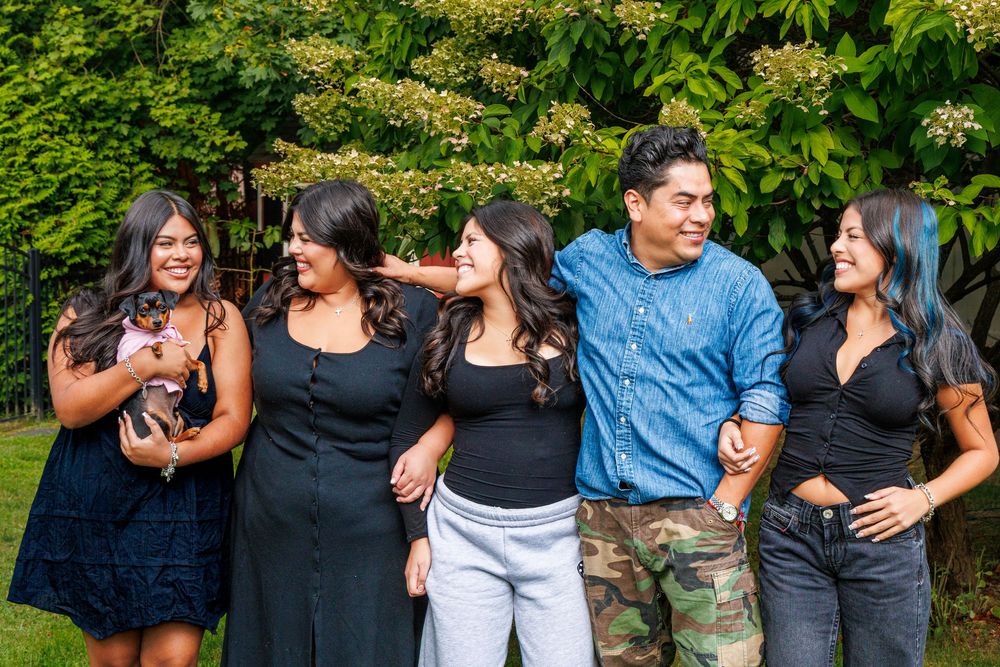 A group poses together outdoors with greenery in the background while holding a small dog.