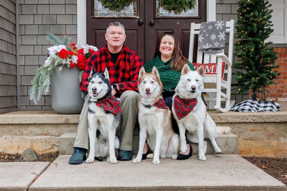 Three Husky dogs wearing matching buffalo plaid bandanas sit on a front porch.