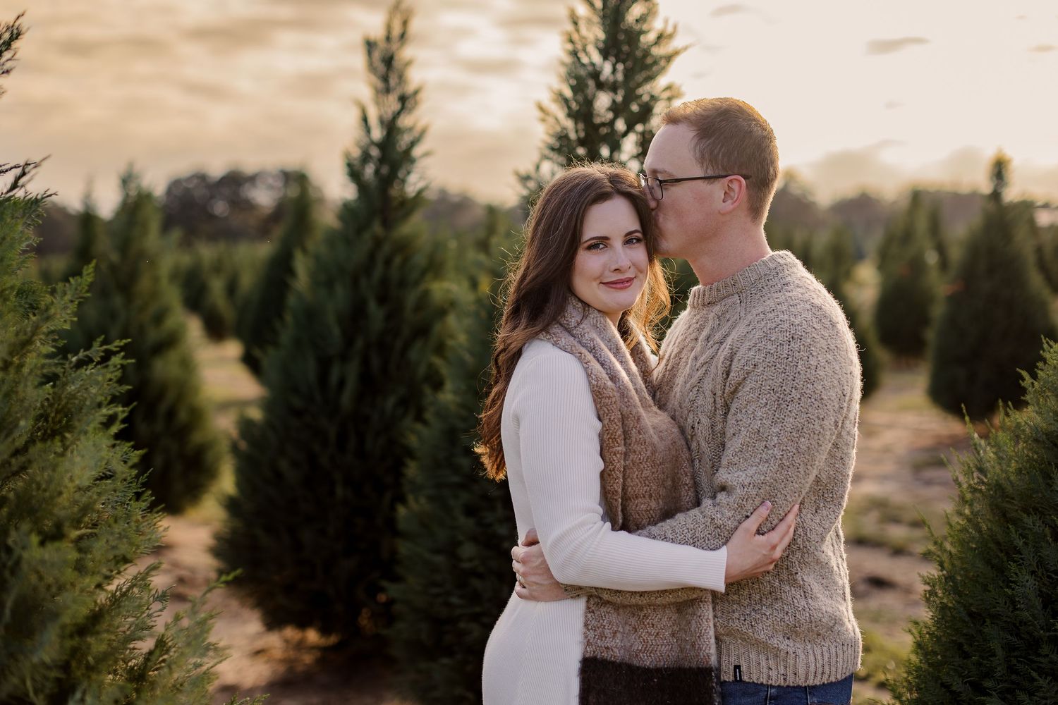 A couple embraces among evergreen trees at sunset on a Christmas tree farm.