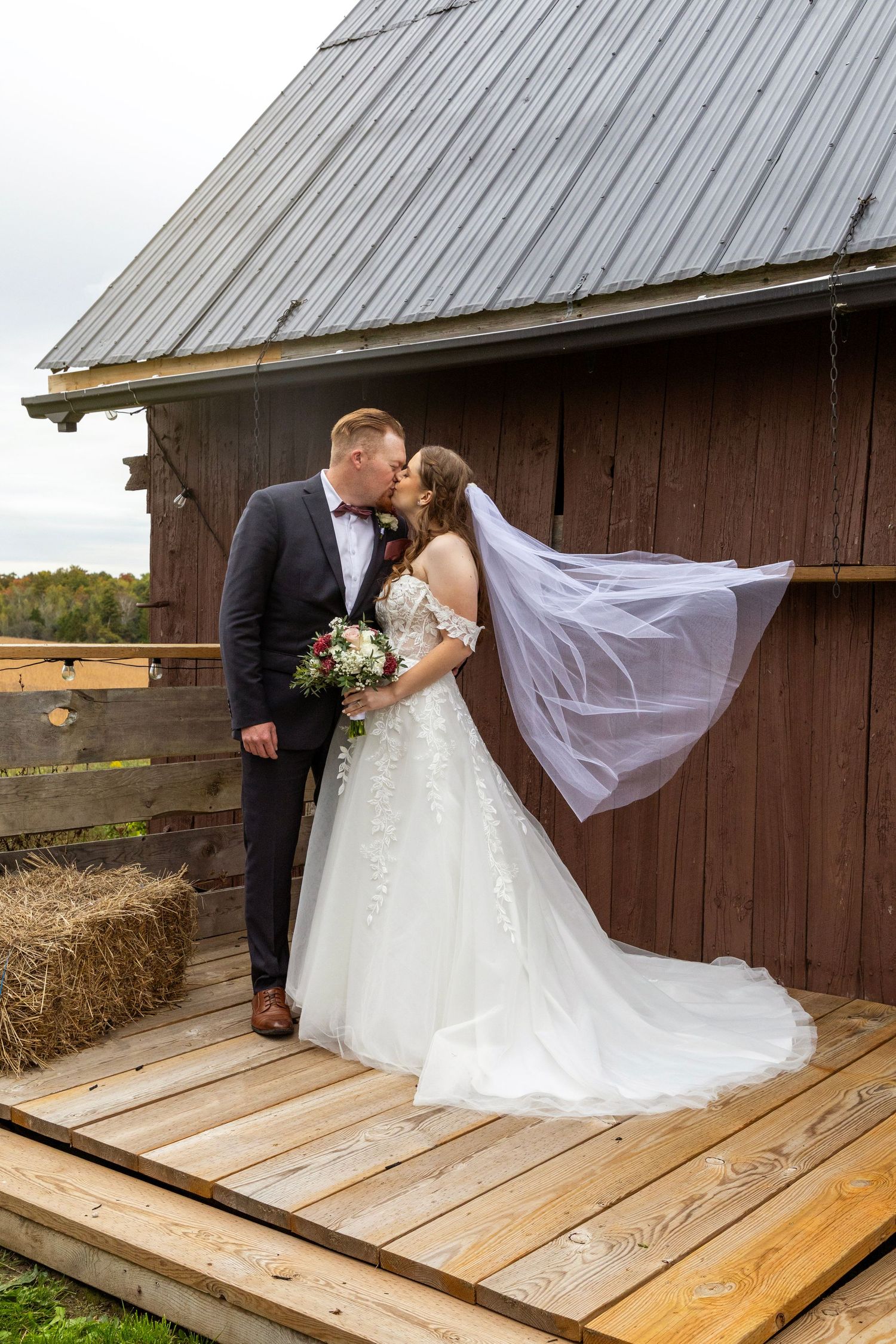 Bride and groom kissing in front of the Lotus Barn in Uxbridge. Bride's veil is effortlessly flowing in the wind