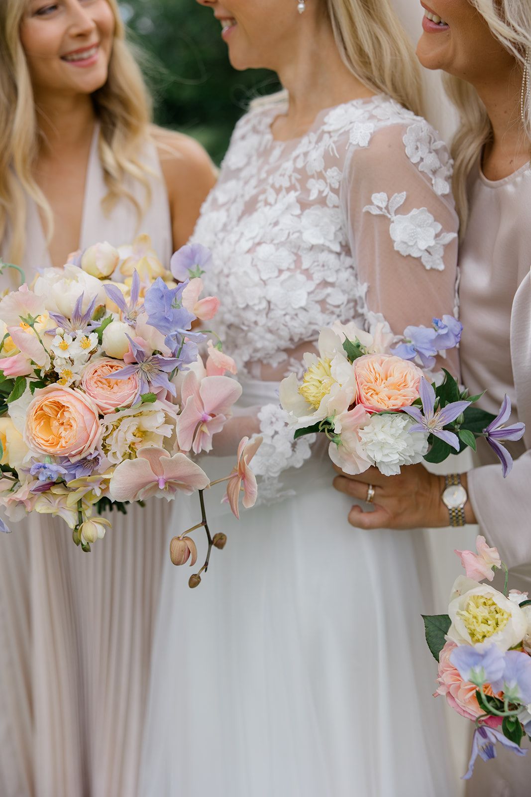 Beautiful pastel wedding bouquets with peach garden roses, lilac blooms and orchids held by bridesmaids in soft neutral dresses.