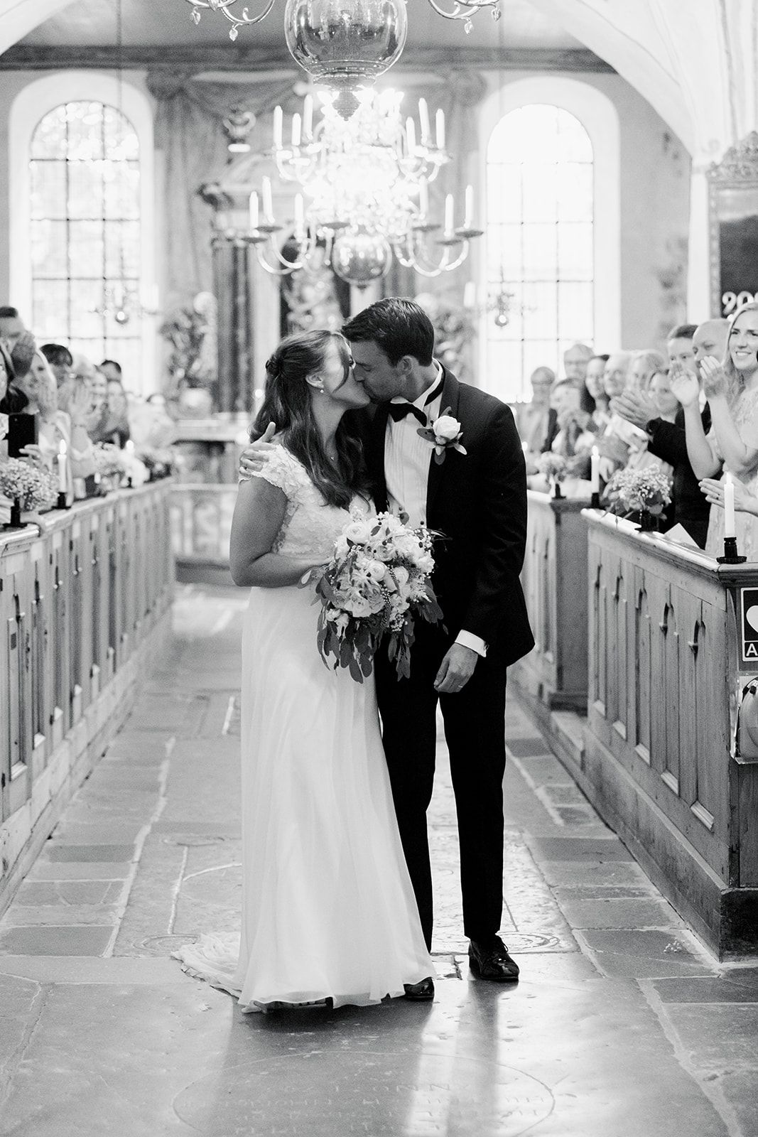 Black and white wedding photos show romantic moments between couple in an elegant church aisle with pews and chandeliers.