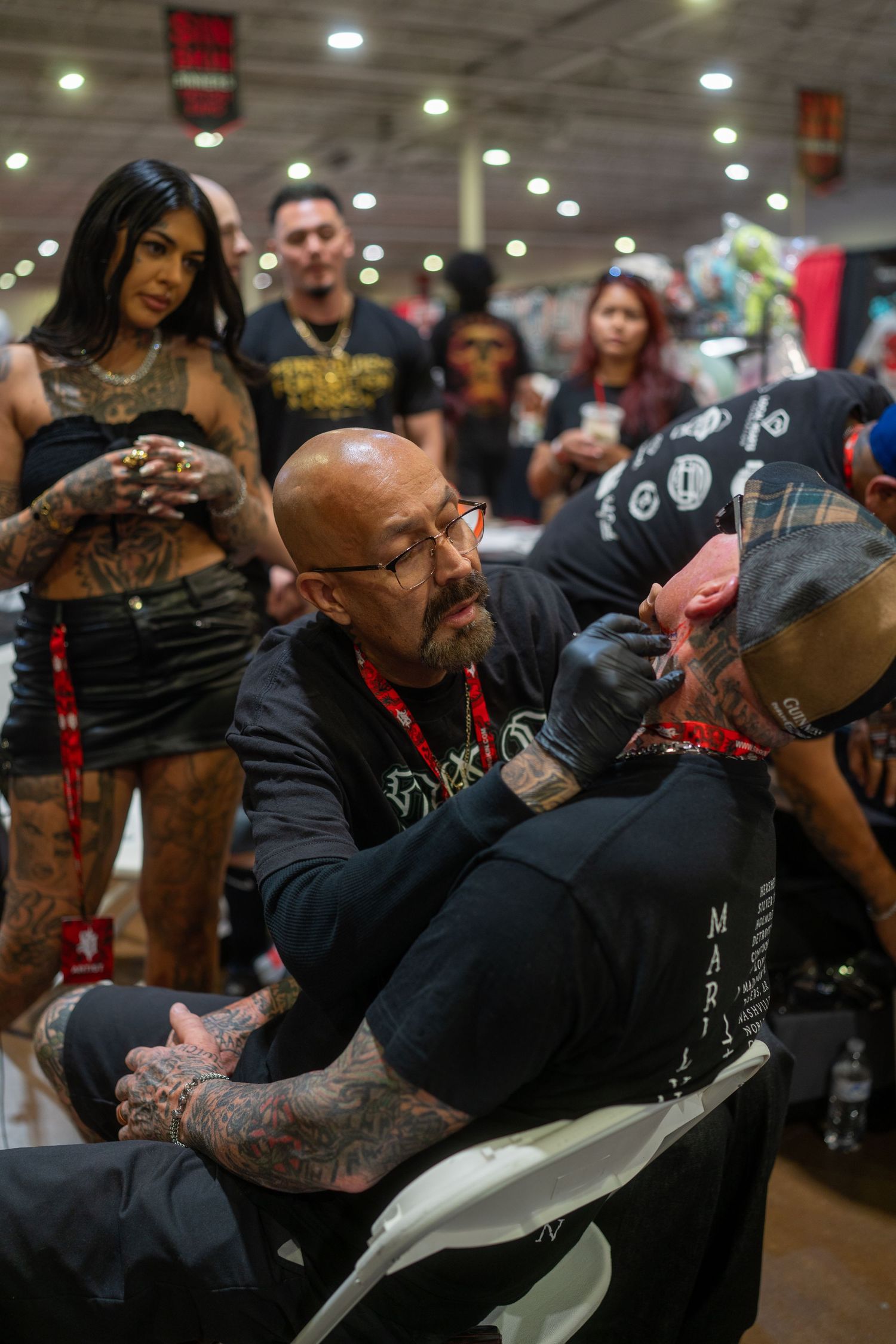 A group of tattoo artists and enthusiasts gather at a busy convention hall with bright overhead lighting.