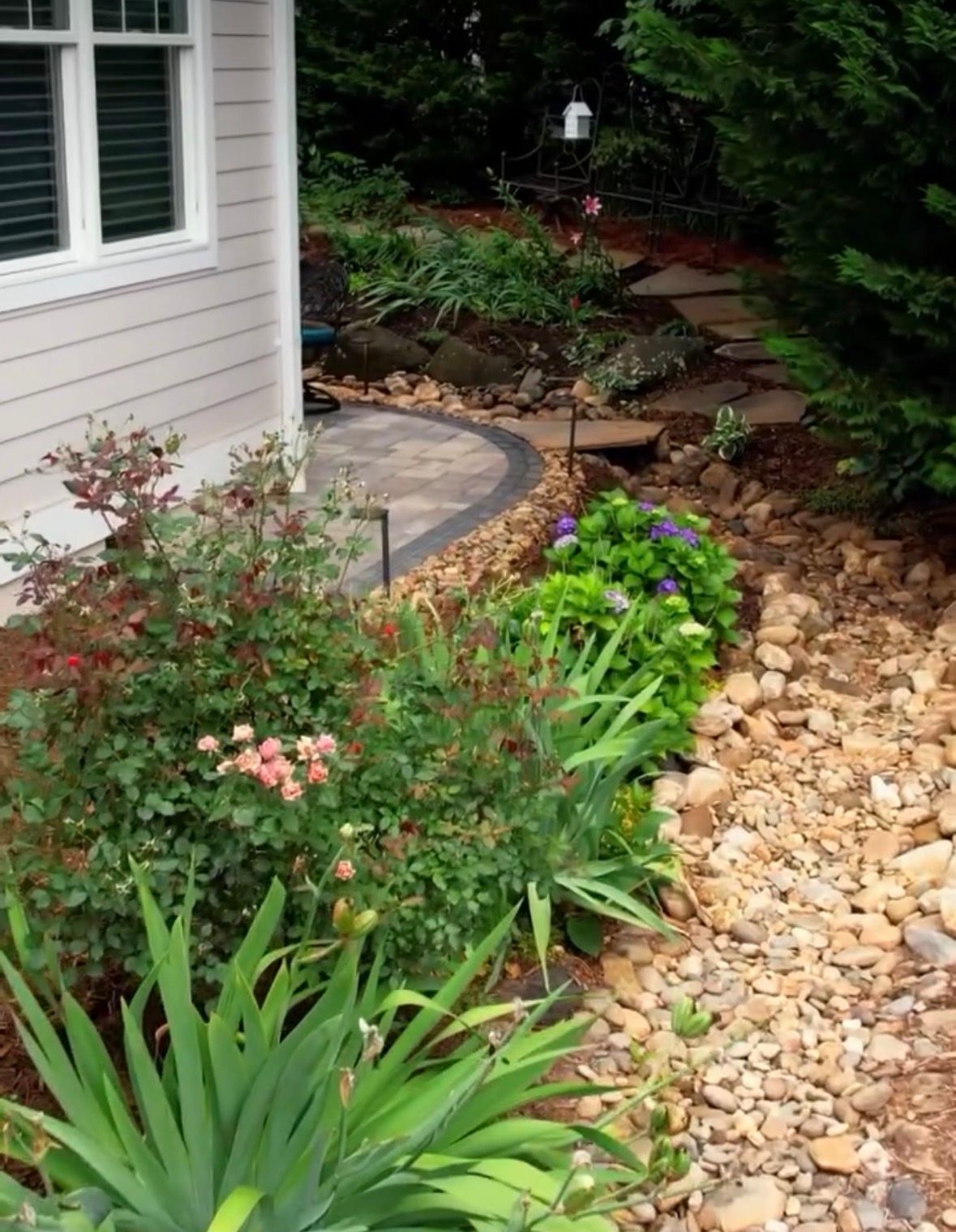 A curved stone pathway leads through a garden with colorful flowers and stacked firewood along a white house exterior.