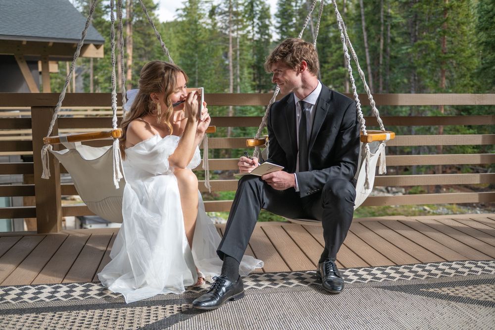 A bride and groom sit in hammock chairs on a wooden deck. They are writing vows in vow books. She has hers across her face and is smiling.