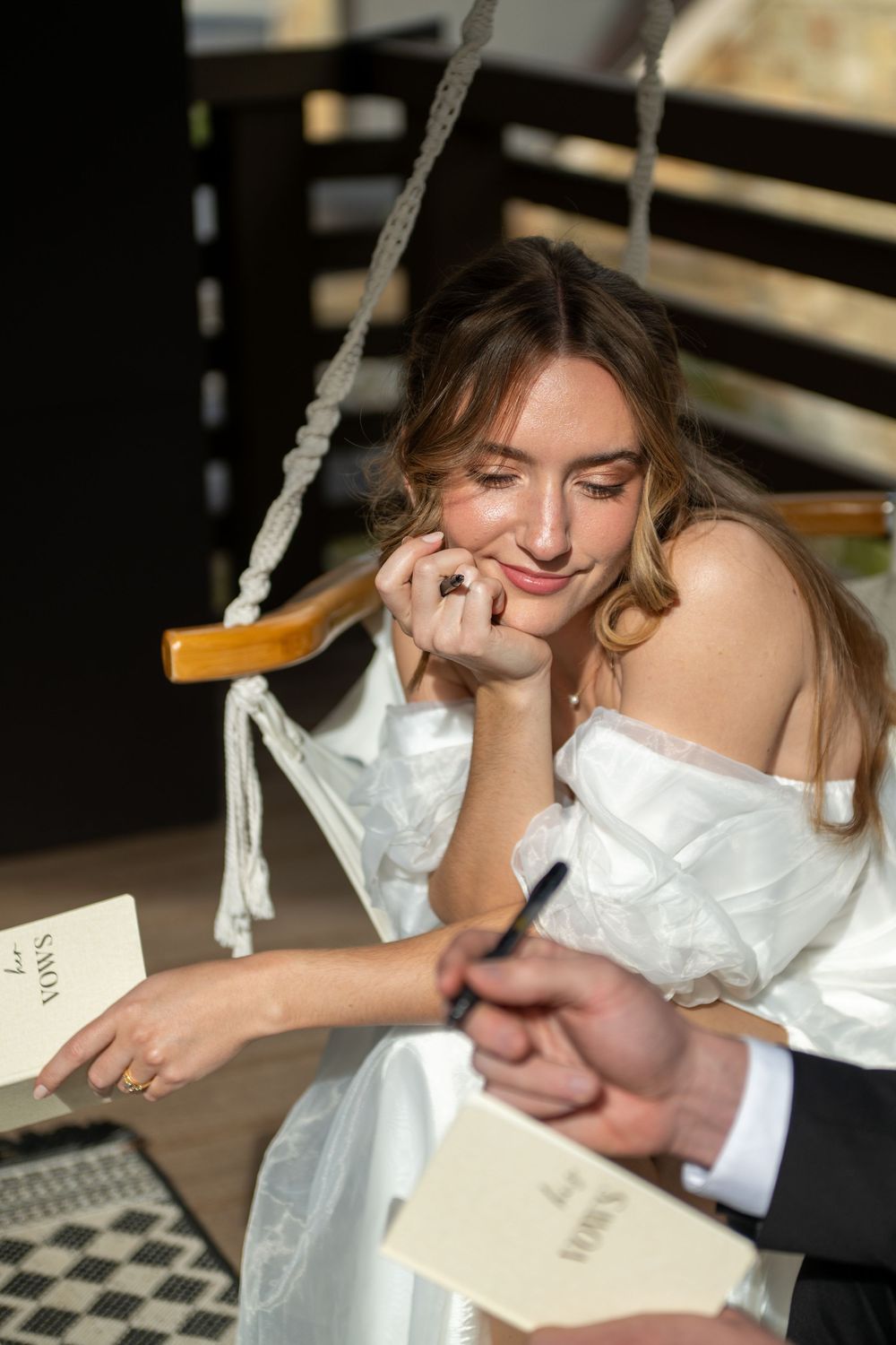 A bride smiles lovingly at her groom's hand as he writes his private vows in a custom vow book. They are sitting in hammock chairs on a deck