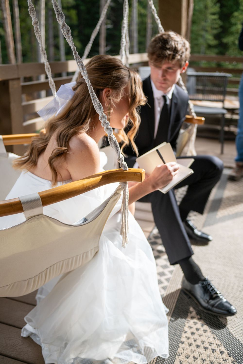 A profile view of a bride and groom in hammock chairs on a wooden deck writing vows to one another. He is looking at her, she is writing.
