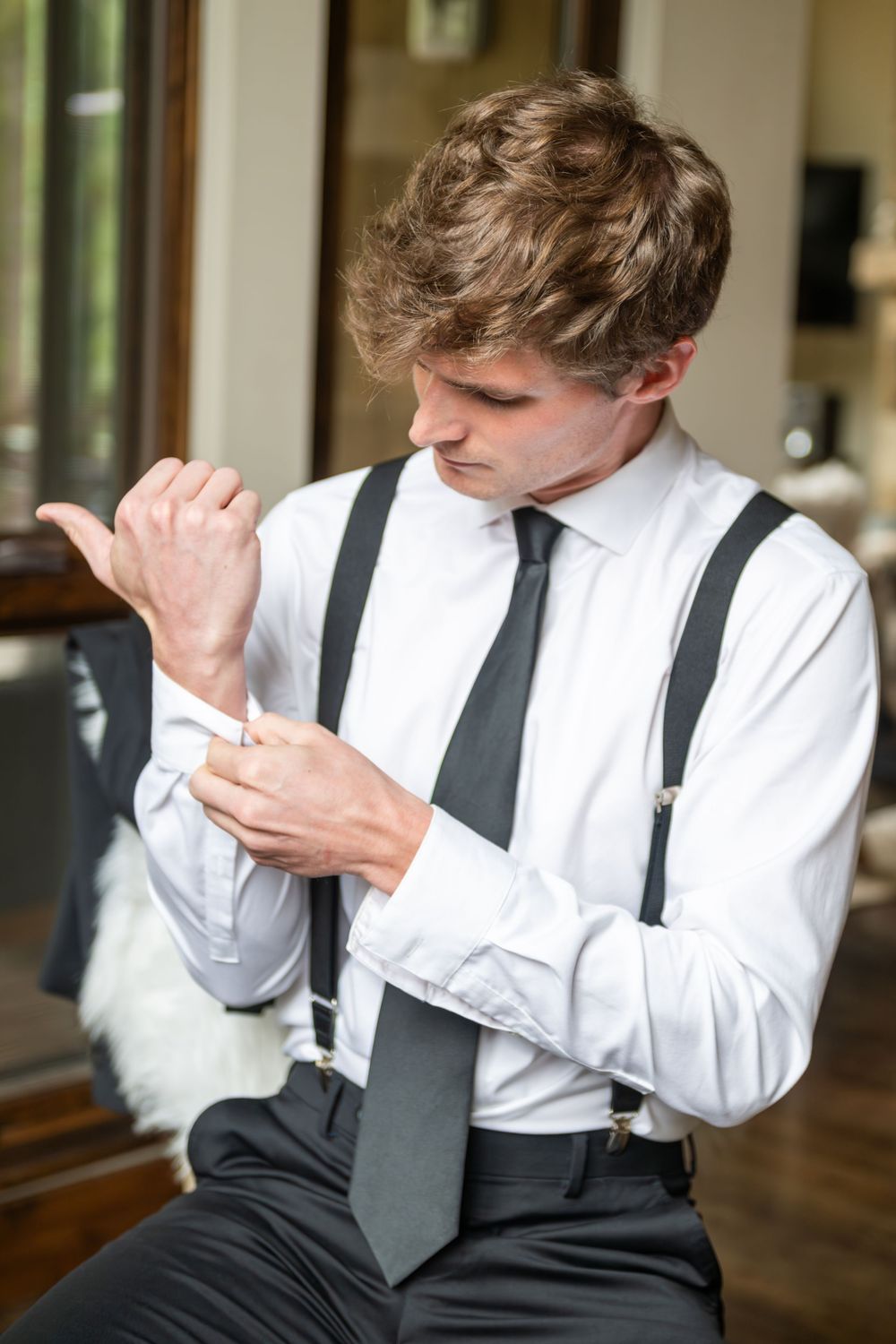 A groom in a clean white shirt and black tie adjusts his cuff links in an upscale vacation rental in natural window light.