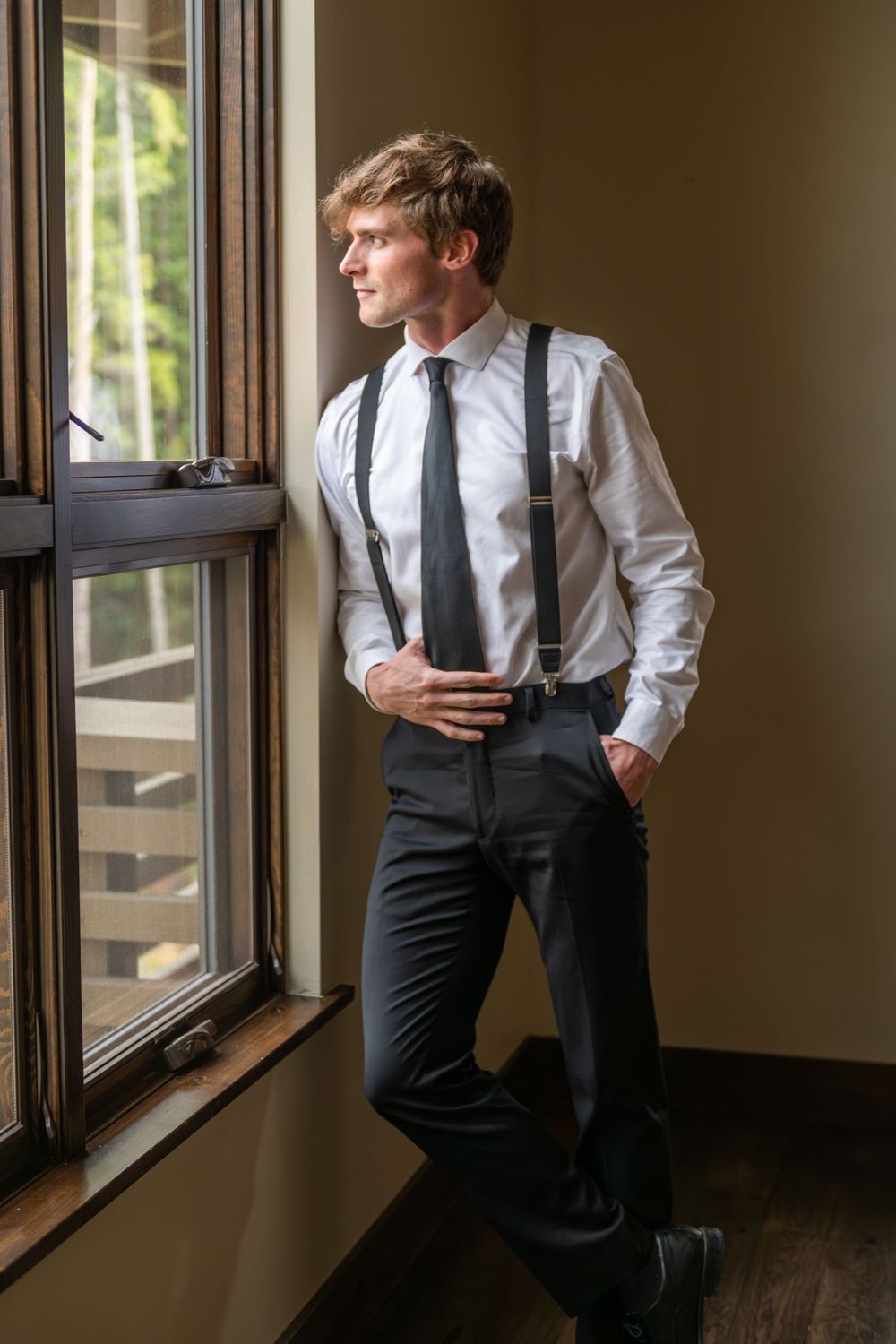 A groom in a clean in white dress shirt and black tie gazes thoughtfully through a window in natural light in an upscale vacation rental.