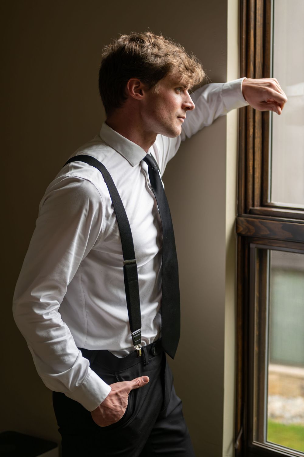 A groom in a clean in white dress shirt and black tie gazes thoughtfully through a window in natural light in an upscale vacation rental.