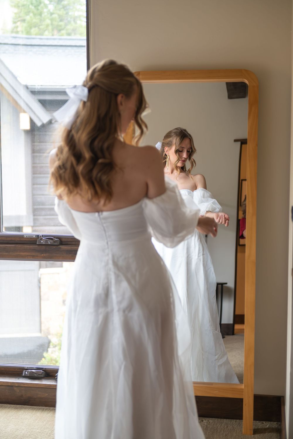 A bride in a white ruffled dress stands in front of mirror while getting ready for her elopement day. Her hair is curled in ringlets.