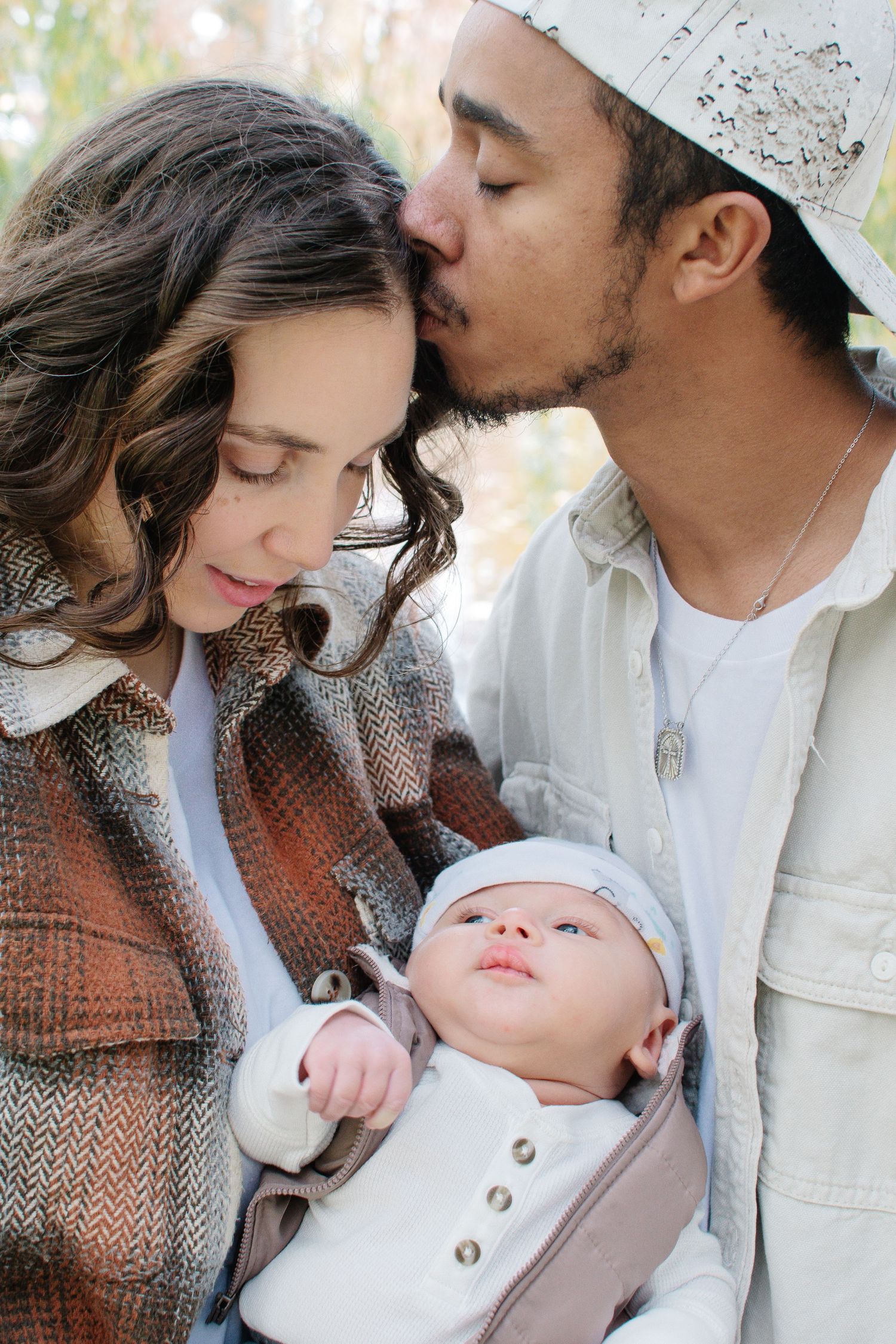 Father kissing newborn while mother looks on during fall family photos at Wright Park in Tacoma.