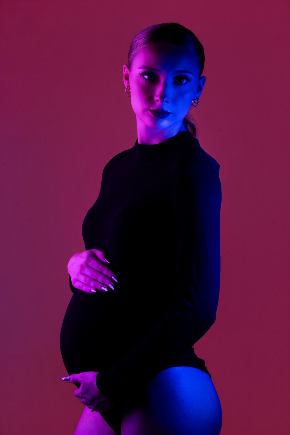 Pregnant woman in black bodysuit illuminated by blue and pink neon studio lighting.