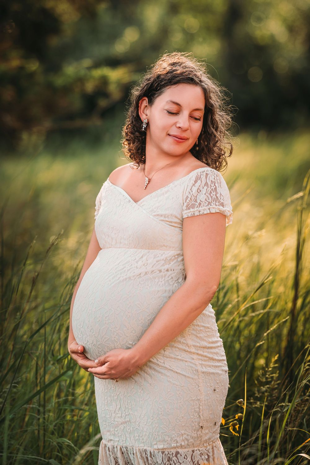 Curly-haired pregnant woman in white lace dress cradling belly in sunlit meadow.