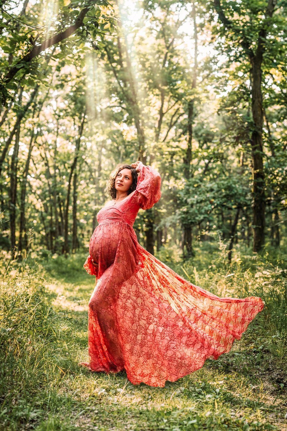 Pregnant woman in flowing red lace dress twirling in sunlit green forest.