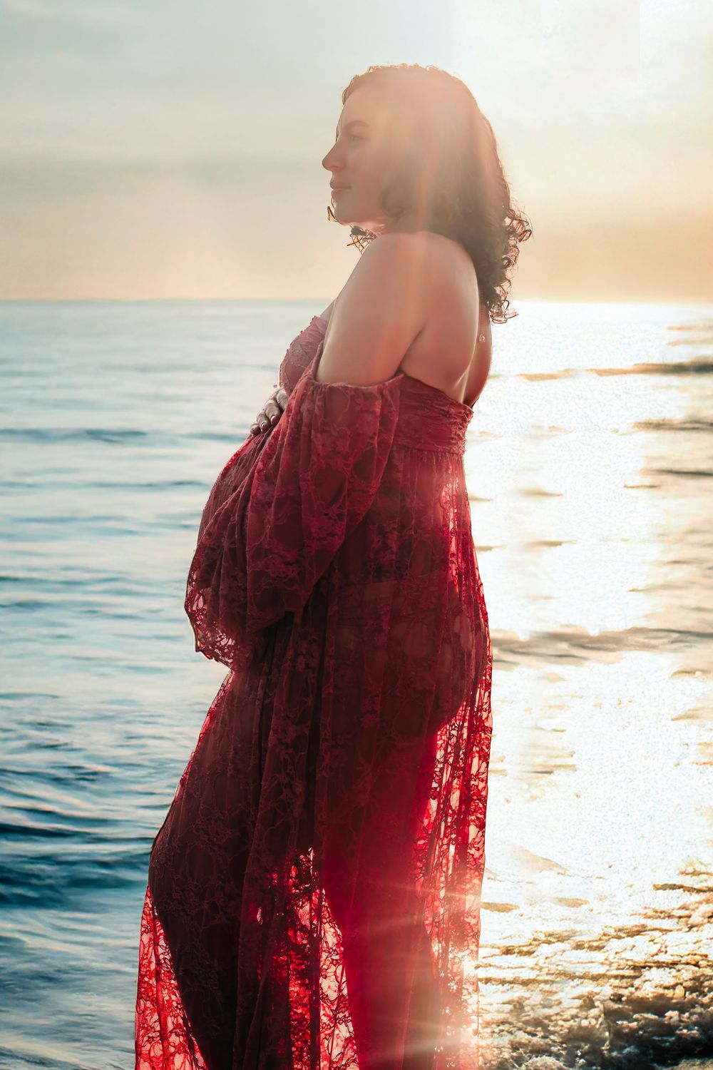Pregnant woman in red lace off-shoulder gown standing by ocean at sunset.