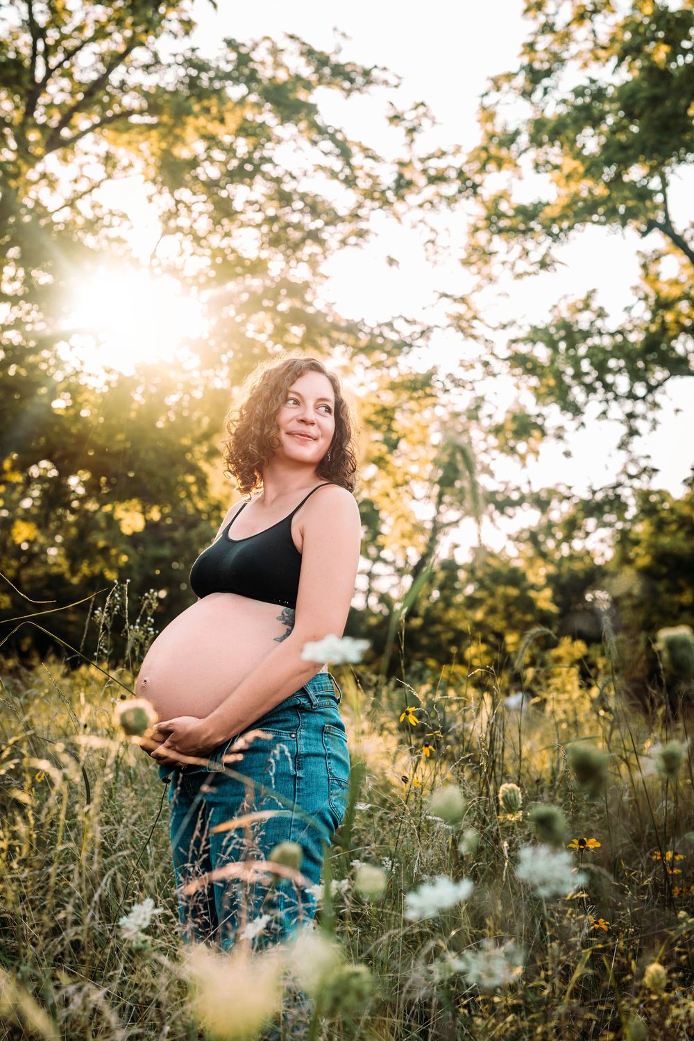Smiling pregnant woman in black top and jeans holding belly in sunny wildflower field.