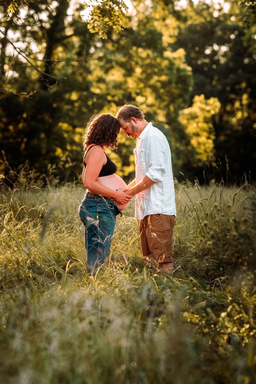 Couple tenderly touching pregnant belly in golden sunlit meadow surrounded by lush green trees.