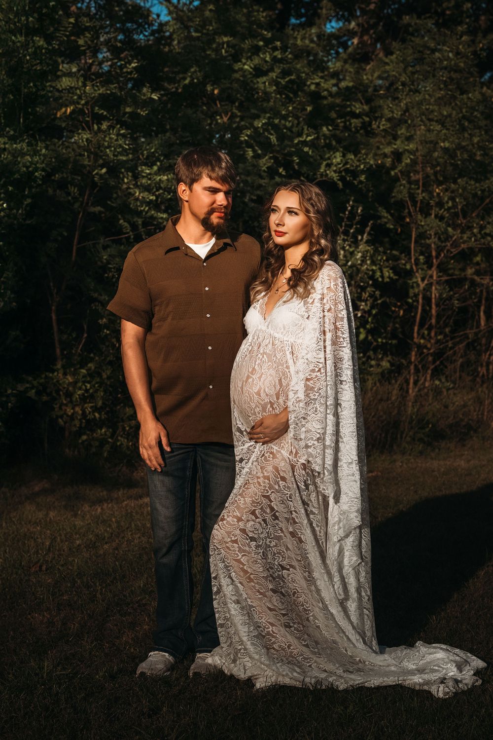 Couple posing outdoors at golden hour, pregnant woman in white lace gown with partner in brown shirt.