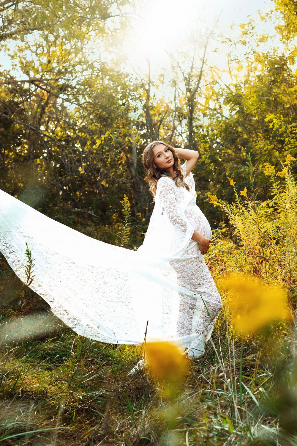 Pregnant woman in flowing white lace maternity gown twirling in sunlit autumn forest.