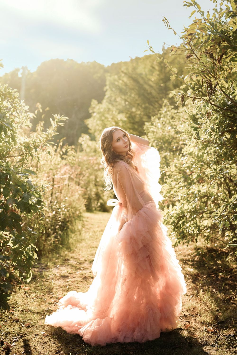 Pregnant woman in elegant pink ruffled gown posing in sunlit garden pathway.
