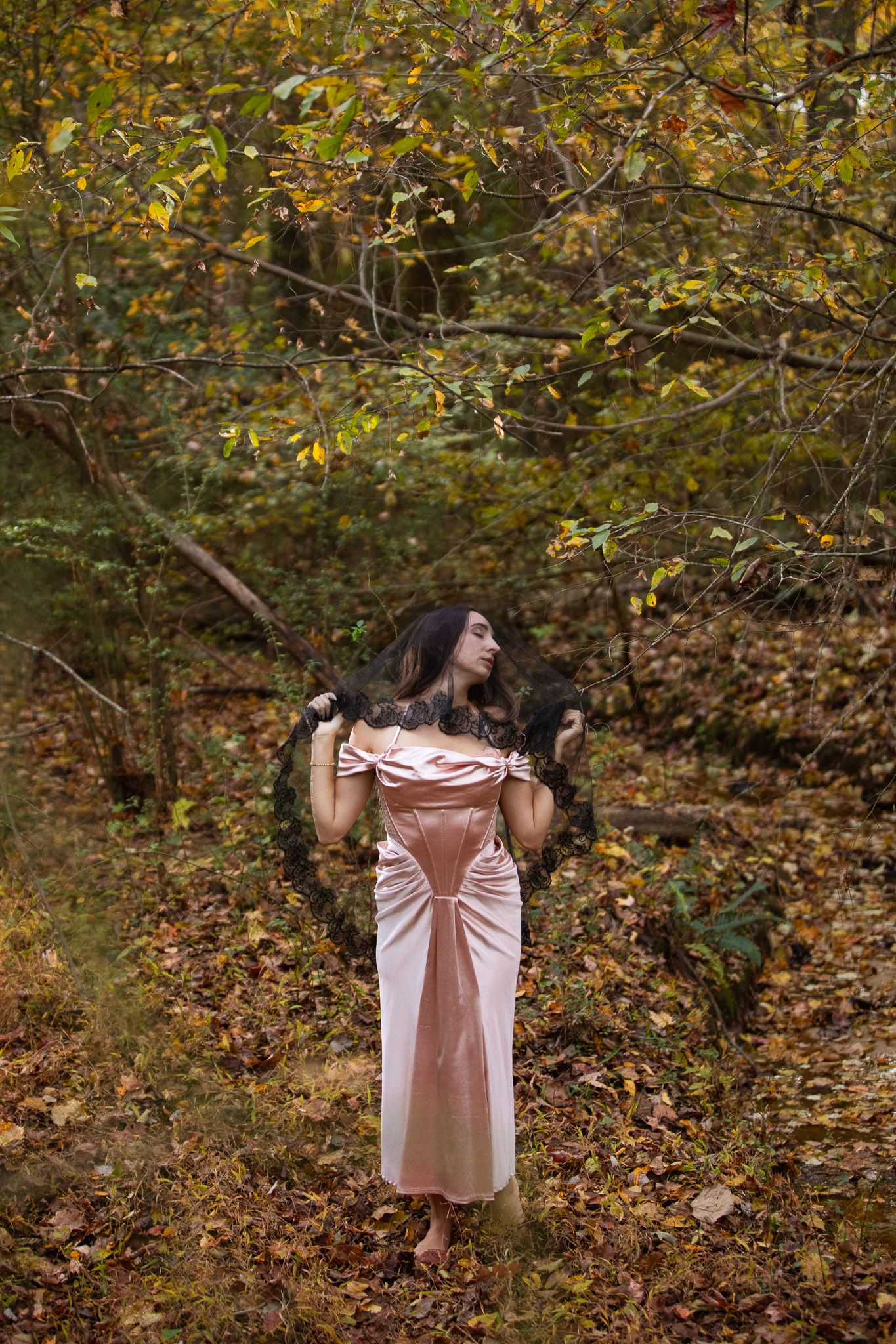 A person in a pink dress stands among autumn trees with fallen leaves creating a dreamy woodland scene.