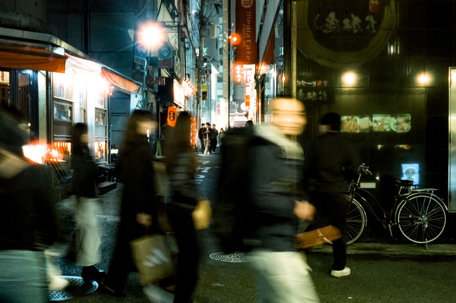 Blurred motion of people walking on a dark city street at night with glowing storefronts and lights in the background.