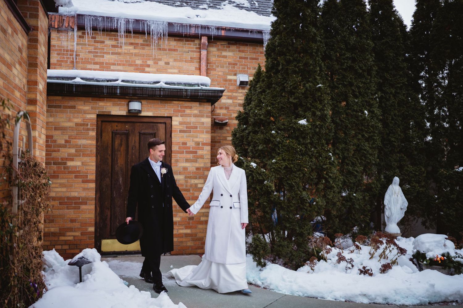 A winter wedding scene outside a brick church with snow on the ground and evergreen trees in the background.