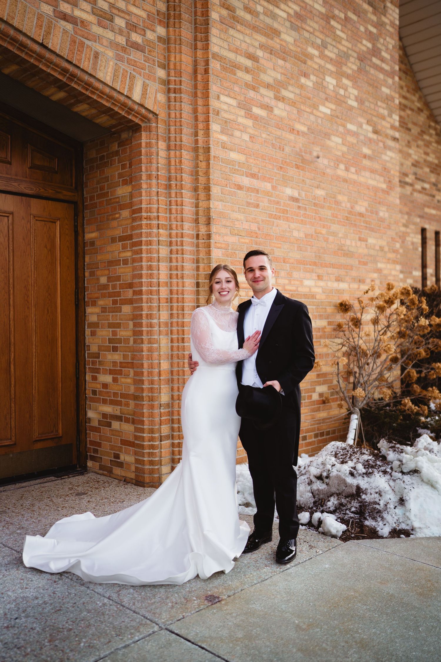 A series of romantic wedding portraits taken against a tan brick church wall with snowy landscaping.