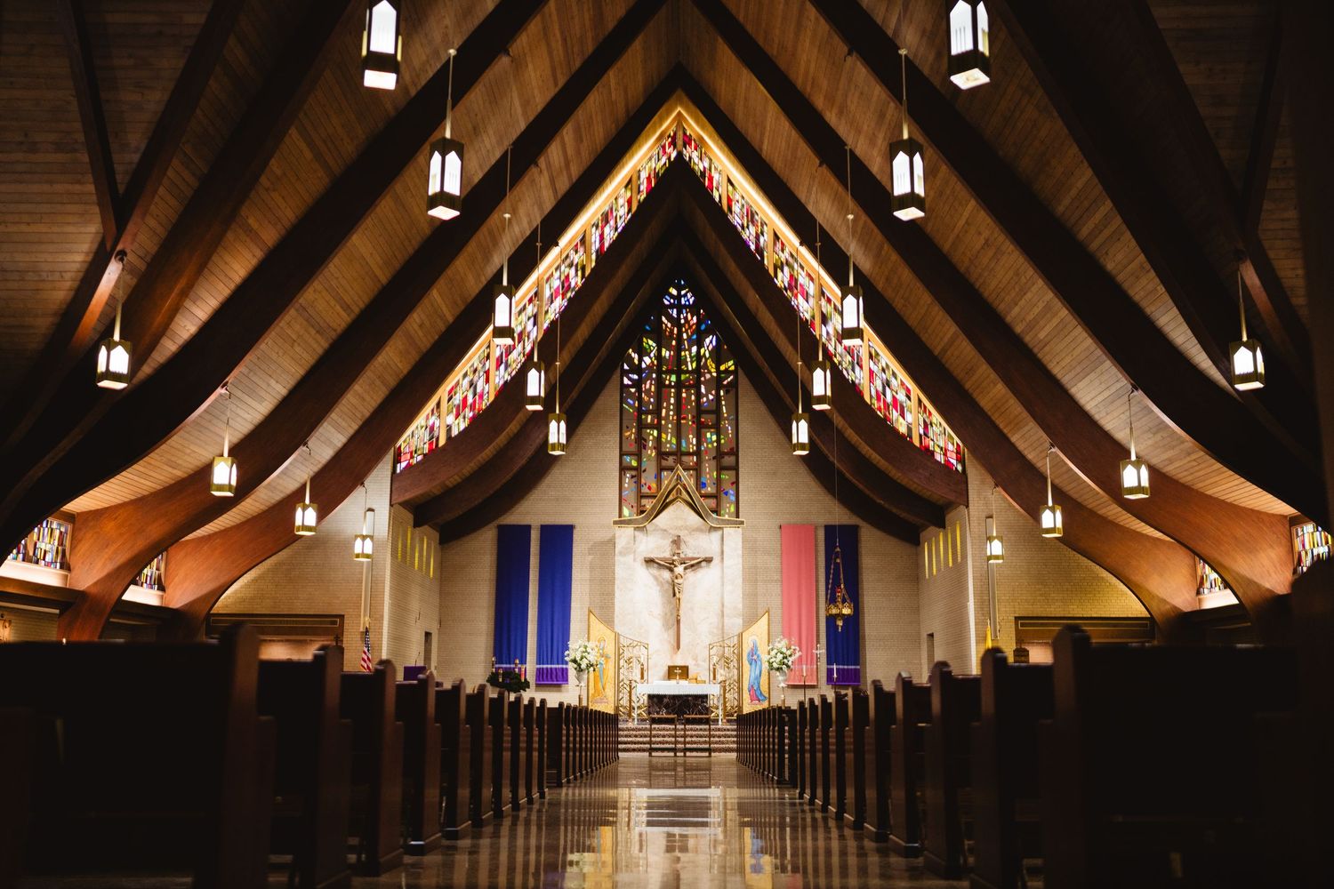 Interior view of a modern church sanctuary with dramatic vaulted wooden ceiling, hanging lights and stained glass altar area.