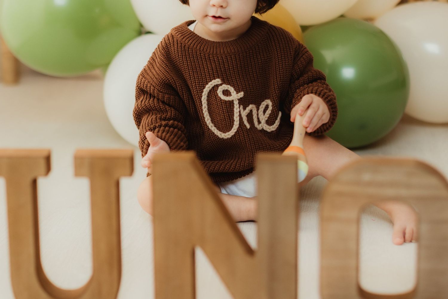 Baby wearing brown sweater sits among green and white balloons at first birthday celebration.
