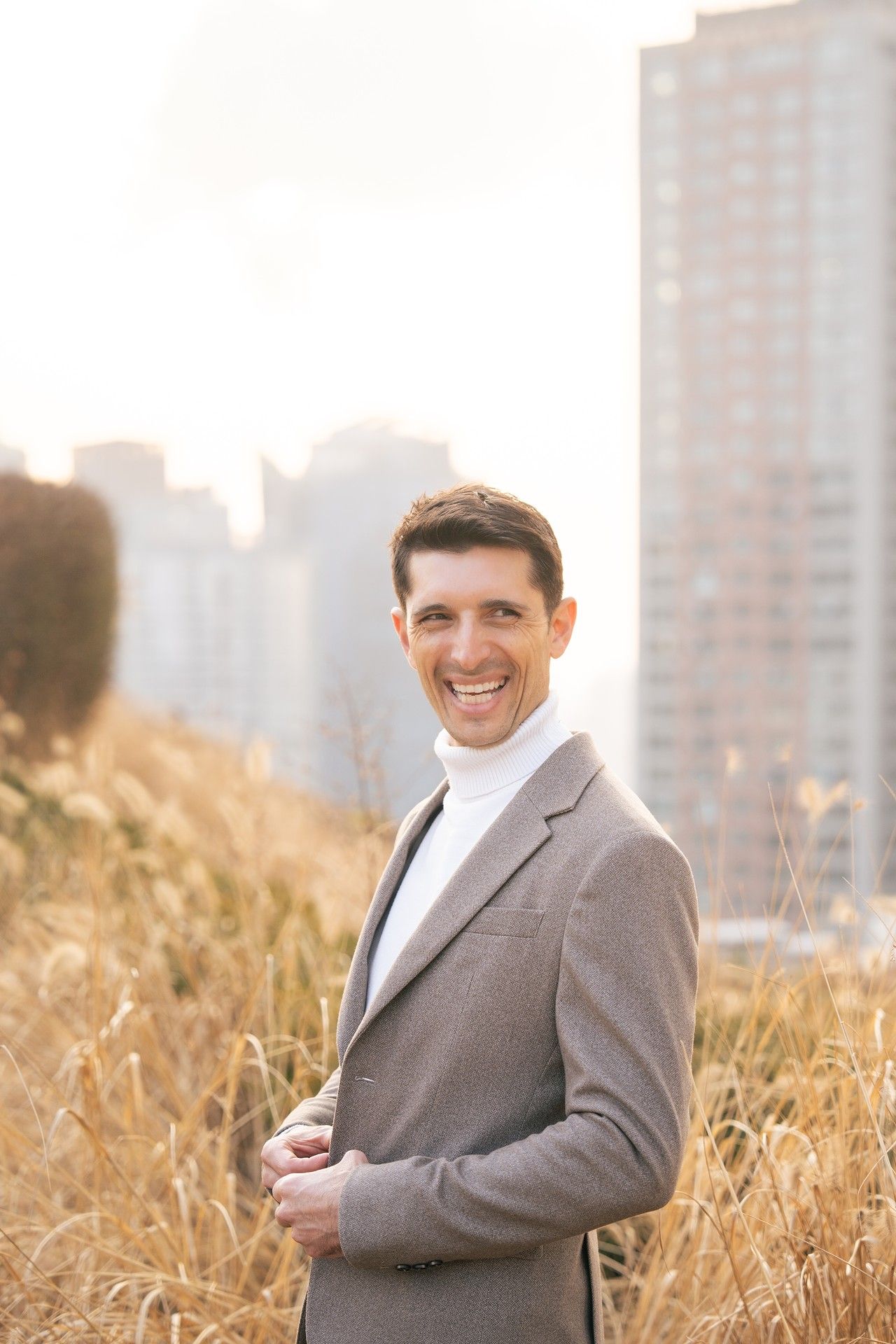 Professional wearing a gray suit jacket poses in a golden wheat field with city buildings in the background at sunset.