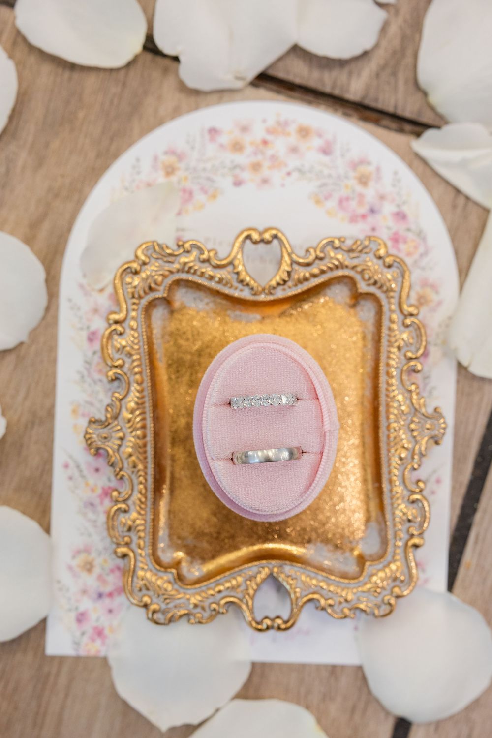 Elegant gold ornate frame holding a diamond engagement ring on pink cushion surrounded by white rose petals.