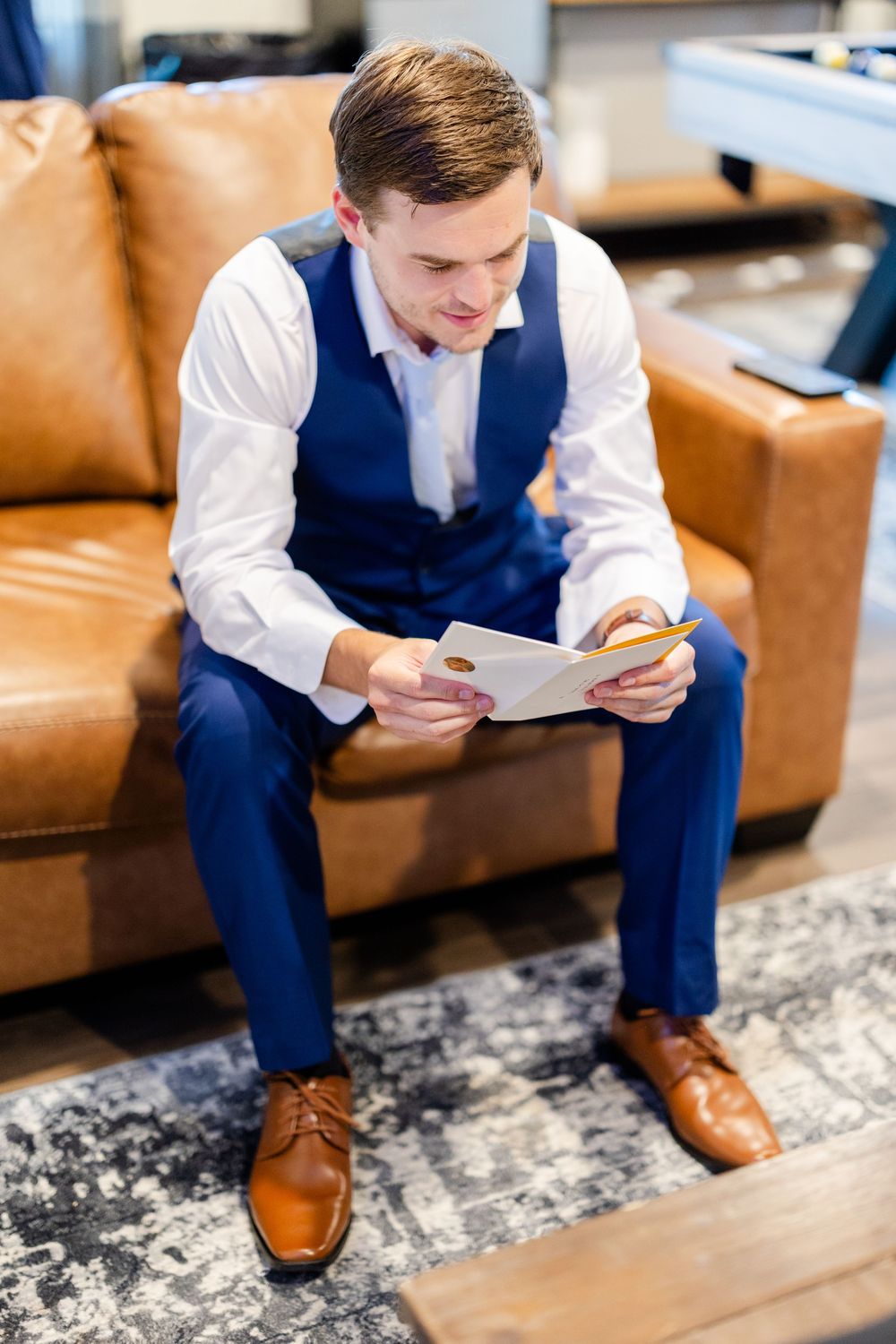 Groom in navy suit vest and brown shoes reads document while sitting on leather couch.
