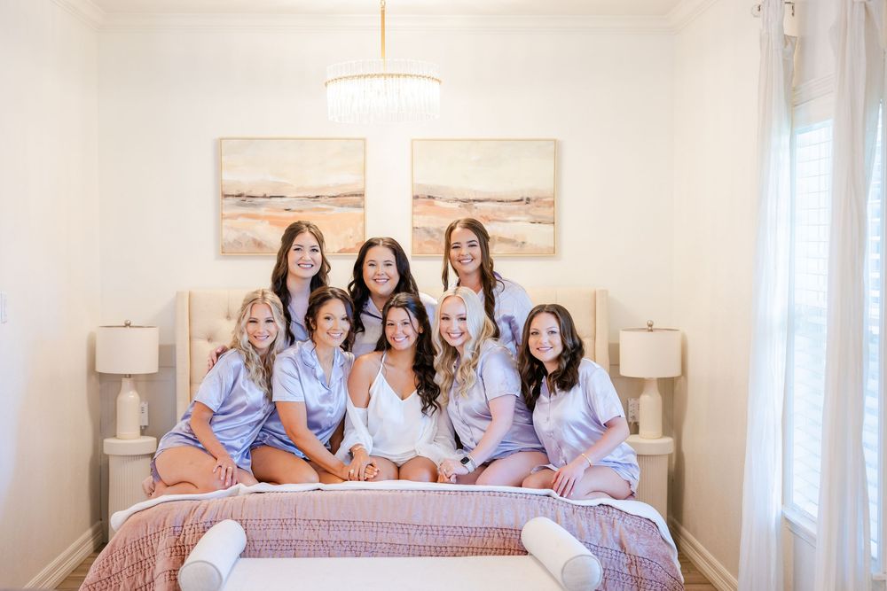 Five bridesmaids wearing matching blue robes sit together on a white bed during wedding preparations.