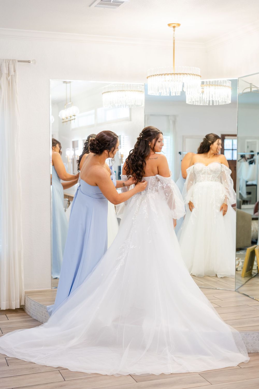 A bridal gown fitting scene showing attendants helping with a flowing white wedding dress in a boutique.