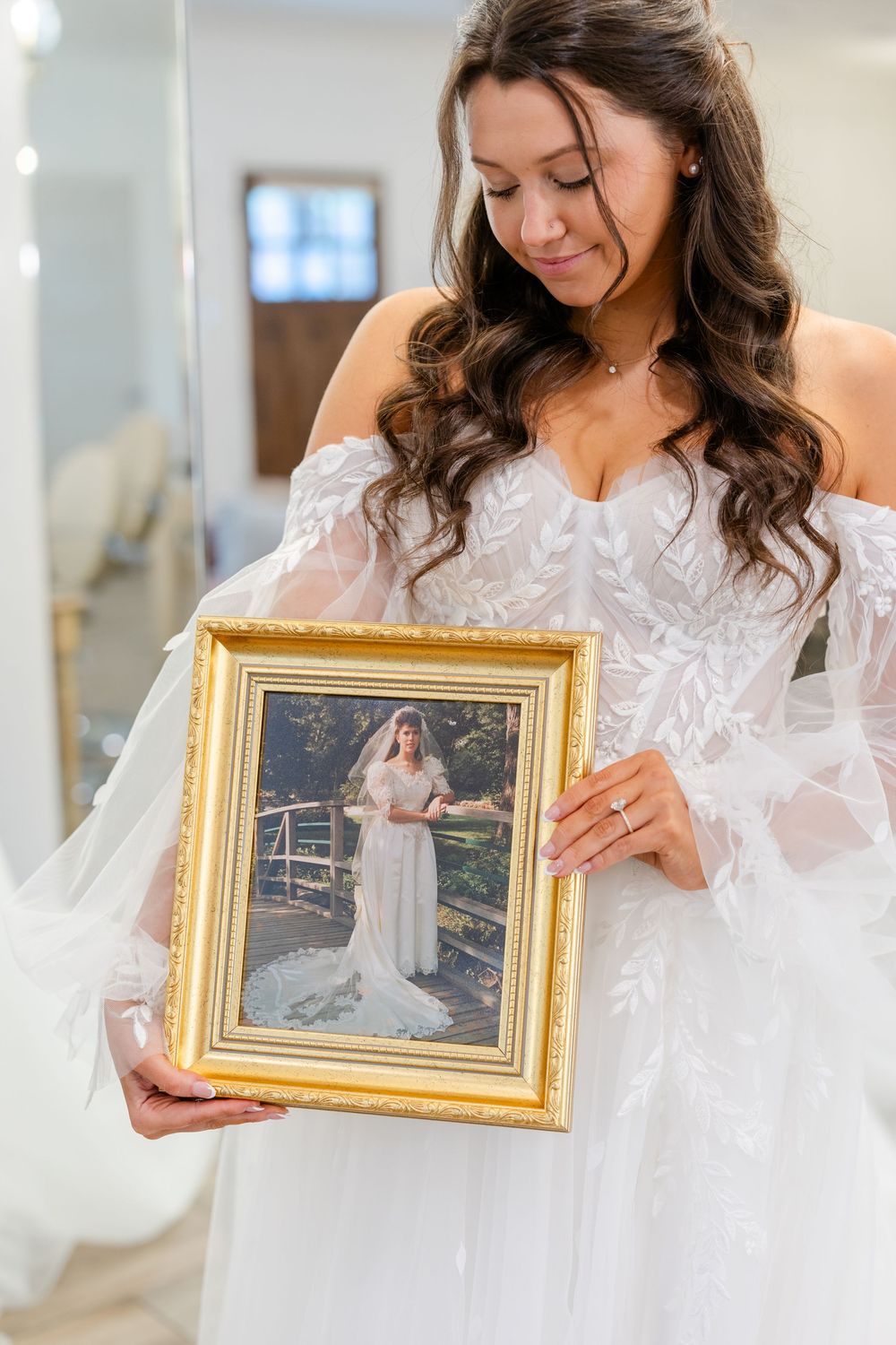 Bride in lace gown holds framed photo of wedding dress fitting.