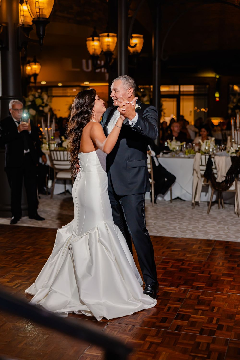 Newlyweds perform their first dance under warm lighting on a parquet dance floor at an indoor reception.
