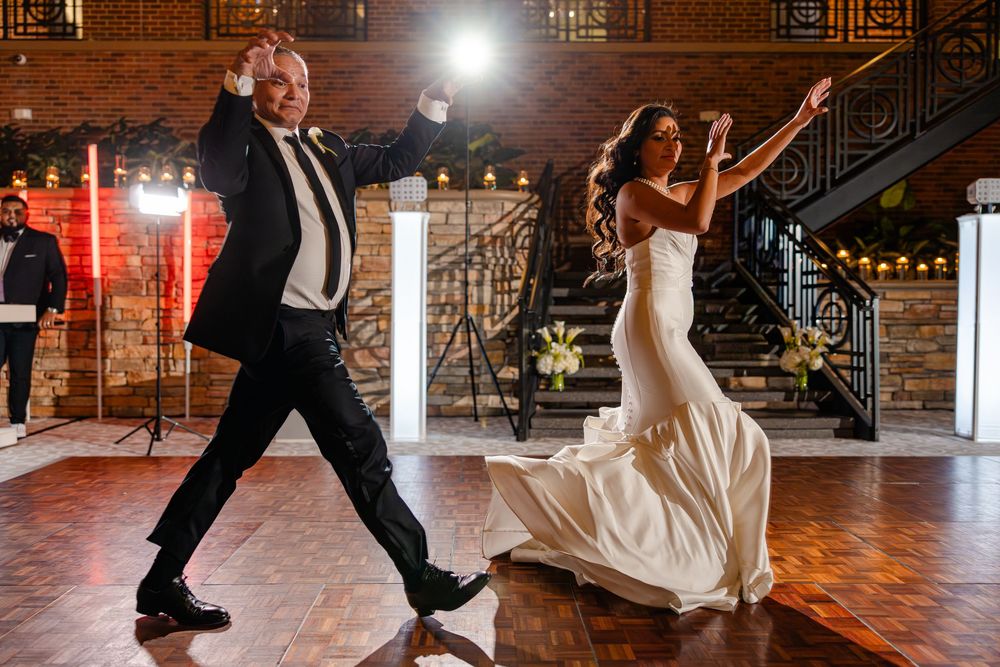 A couple performs a fun dance move at a wedding reception in a rustic brick-walled venue with warm lighting.
