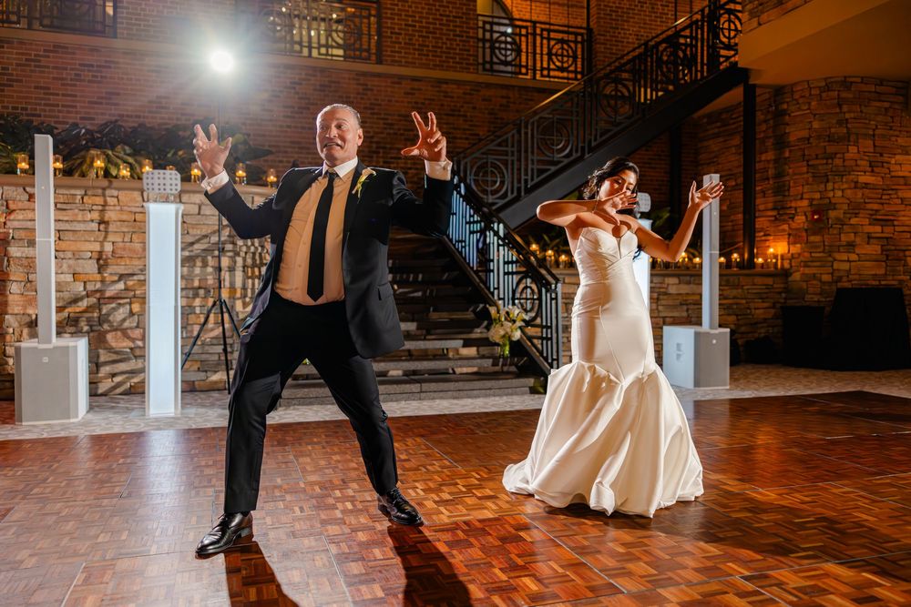 Wedding couple performs an energetic dance move in an industrial-style venue with brick walls.
