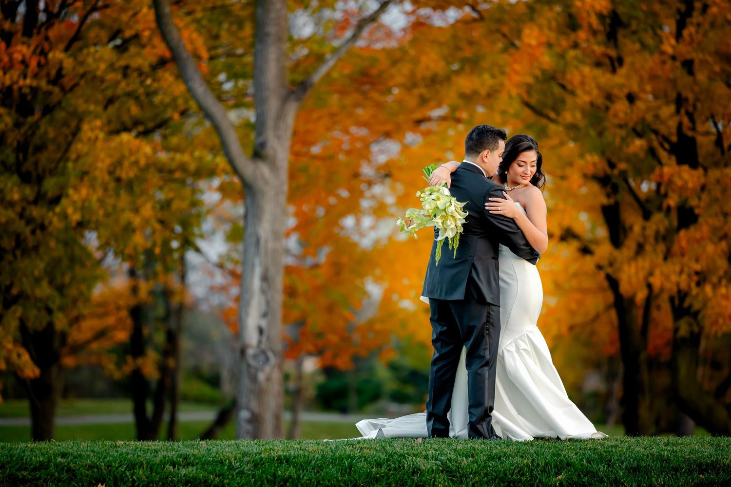 A couple in wedding attire embraces among vibrant orange autumn trees in a park setting.