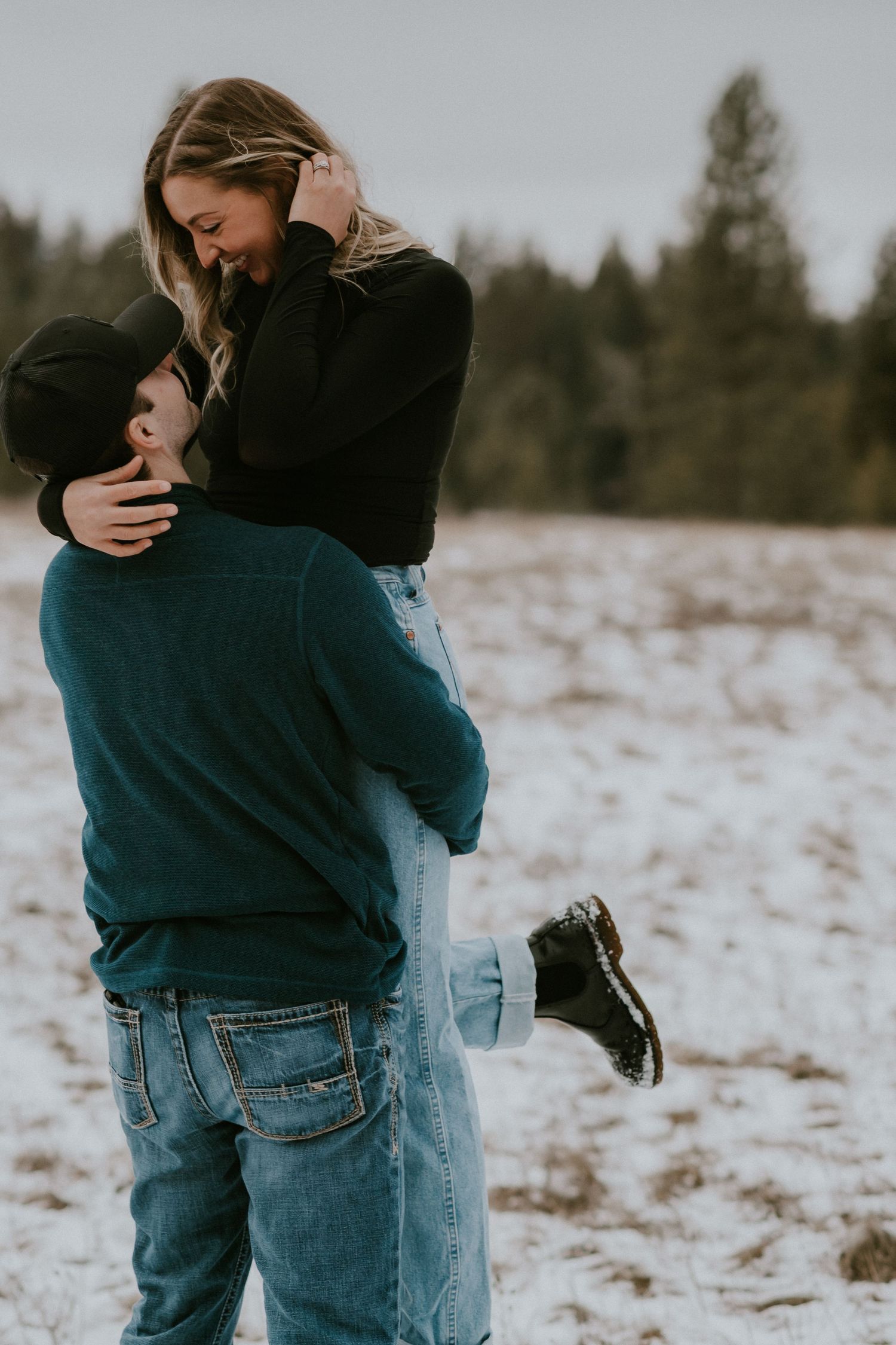 A couple embraces in a romantic winter scene with snow-covered ground and evergreen trees in the background.