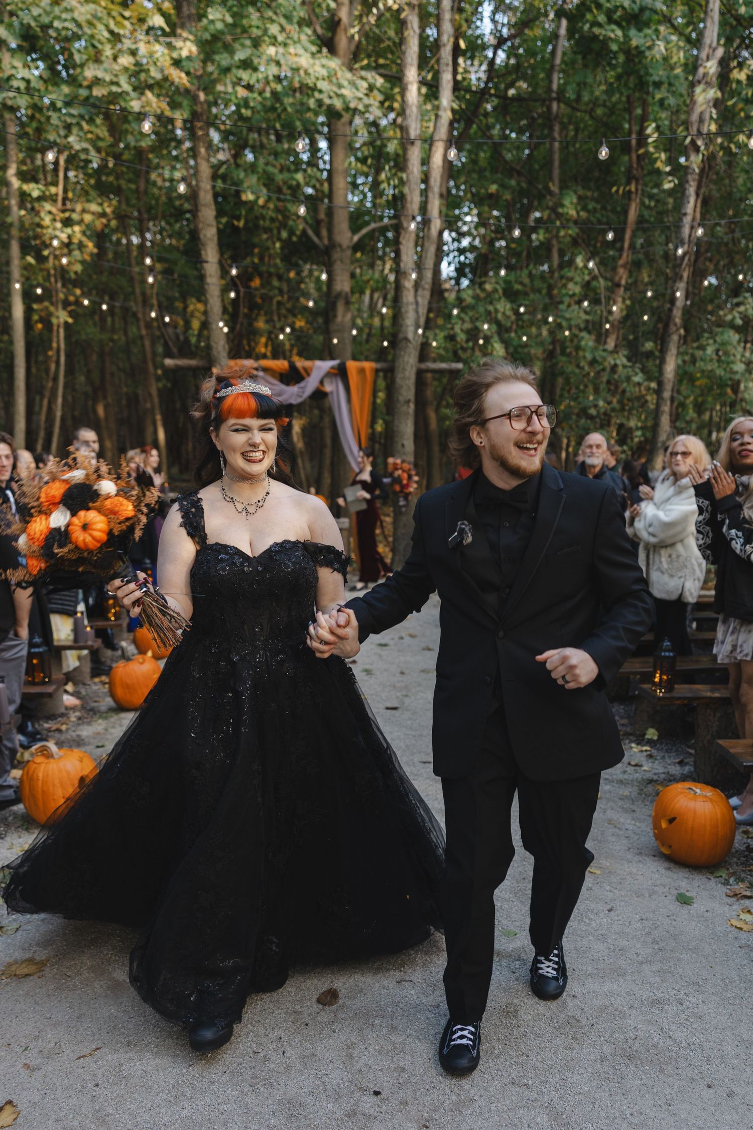 Wedding guests dance and celebrate at an outdoor reception with orange pumpkin decorations and trees in the background.